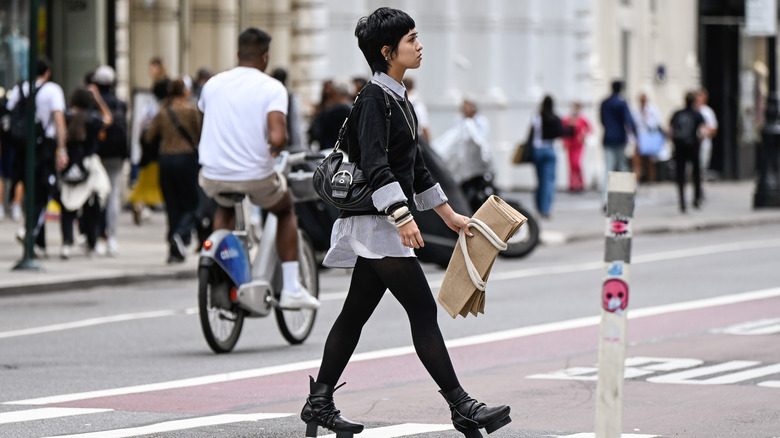 Une personne traverse la rue dans des leggings noirs et un gilet de pull noir sur un chemisier blanc