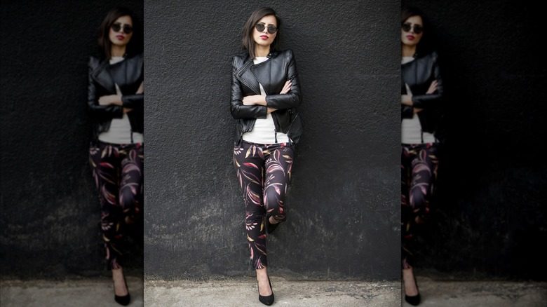 Une femme se tient contre un mur dans des lunettes de soleil fraîches, des talons, des leggings à motifs et une veste en cuir