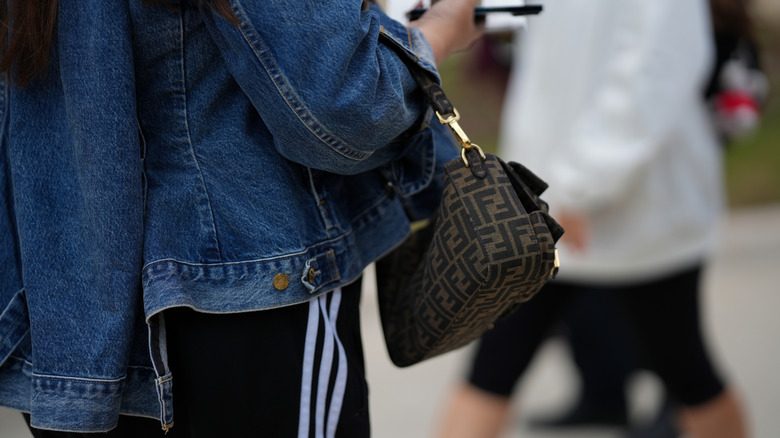Une personne tenant un pantalon de piste, une veste en jean et accesorisant avec un sac Fendi