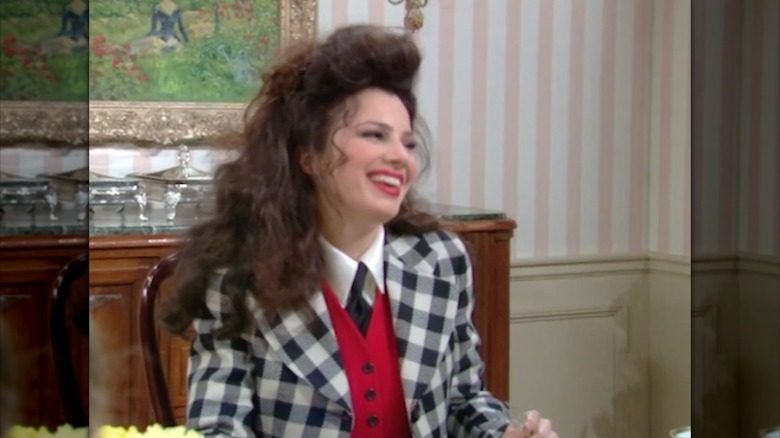 Fran Fine rit à la table du petit-déjeuner en portant un gilet rouge, un blazer à carreaux noir et blanc, un col blanc et une cravate noire