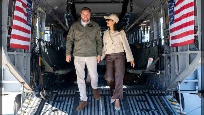 Usha Vance marche main dans la main avec JD Vance sur un porte-avions. Elle porte un trench-coat court beige, un pantalon en cuir marron et des talons blancs à bout pointu.