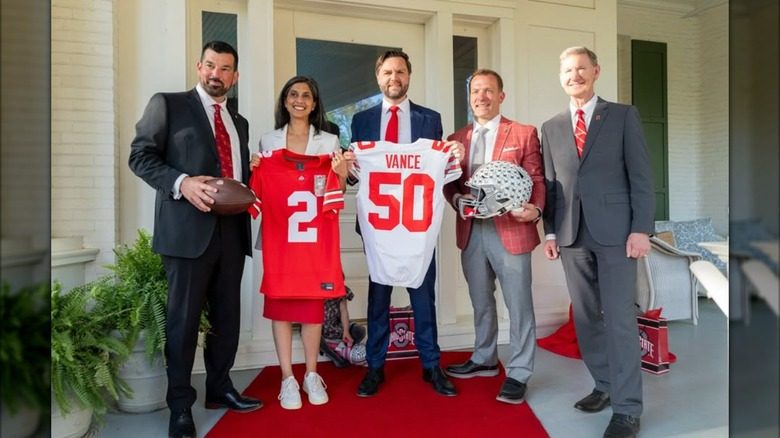Usha Vance brandit un maillot de football rouge à côté de son mari JD Vance et d'autres hommes. Elle porte un blazer blanc, une longue jupe rouge et de grosses baskets blanches.
