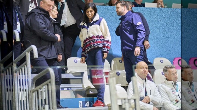 Usha Vance et JD Vance parlent aux agents de sécurité aux Jeux olympiques d'hiver de 2026. Usha porte des baskets rouges à rayures blanches, un pantalon bleu marine et une veste en laine blanche.