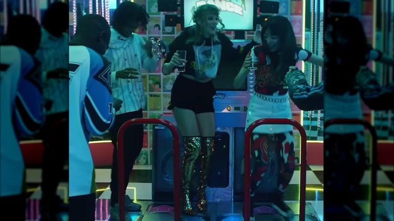 Taylor Swift joue à un jeu de danse dans une salle d'arcade. Elle porte un sweat à capuche graphique court, un short de survêtement noir et une paire de cuissardes à paillettes dorées.