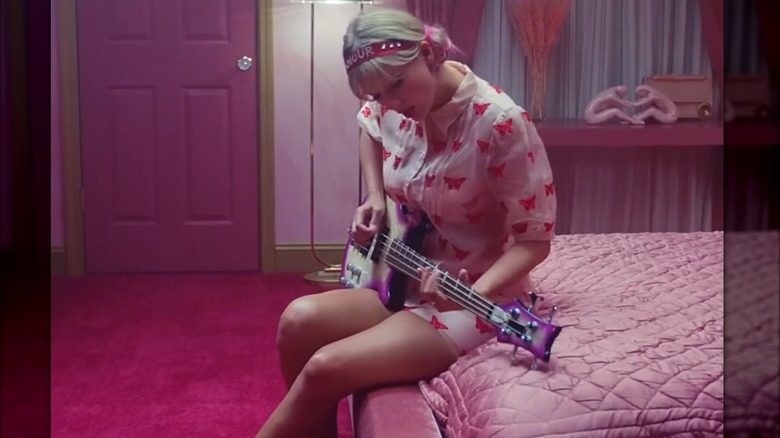 Swift est assis sur un matelas rose et joue de la guitare rose. Elle porte une robe boutonnée rose clair recouverte de papillons rose vif, un bandeau en acrylique rose et des rubans roses pour ramener ses cheveux en deux chignons bas.