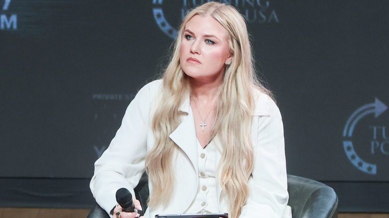 Erika Kirk est assise à un événement Turning Point USA. Cette image montre ses extensions blondes ultra longues. C'est une femme à la peau claire et porte une veste de costume blanche et un gilet assorti.