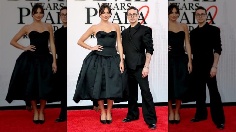 Paige DeSorbo pose avec Christian Siriano. DeSorbo est une jeune femme à la peau claire et aux cheveux foncés. Elle porte une robe noire sans manches avec une jupe trapèze taille basse. Christian Siriano est un homme à la peau pâle et aux cheveux noirs courts. Il porte un costume noir avec des manches remontées sur les bras.