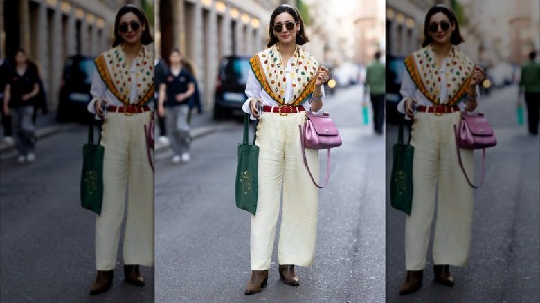 Une femme pose dans un chemisier boutonné, un pantalon crème et une écharpe à motifs.