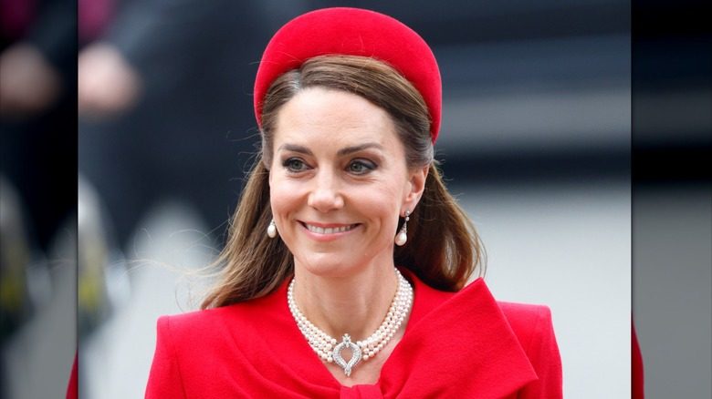Kate Middleton sourit en marchant dans la rue dans une tenue rouge et un tour de cou en perles autour du cou
