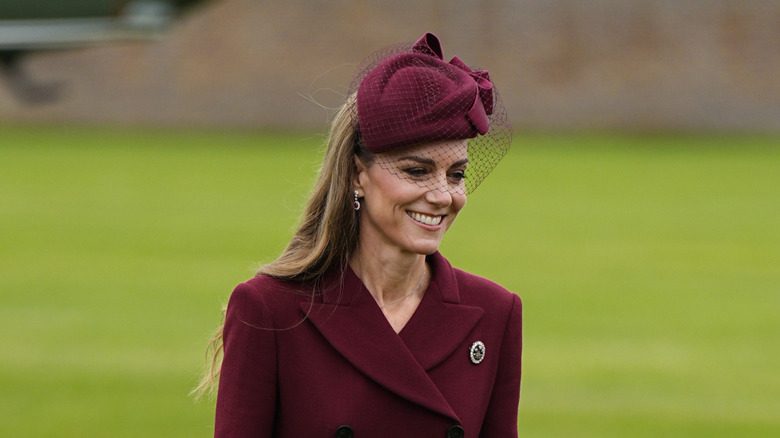 Kate Middleton sourit en marchant dehors, portant une broche sur son manteau canneberge