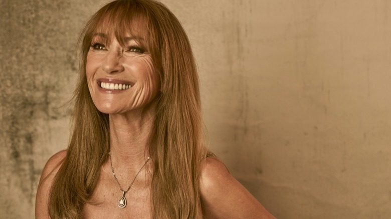 Jane Seymour souriante et portant un collier pendentif sur fond marron.