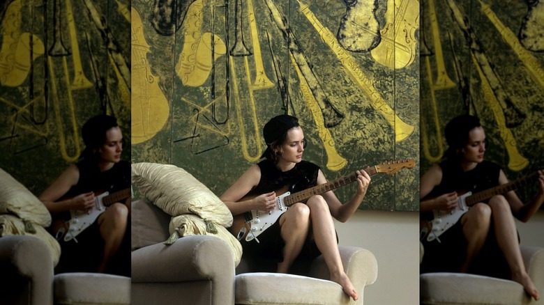 Winona Ryder joue de la guitare sur une chaise devant une œuvre d'art colorée.