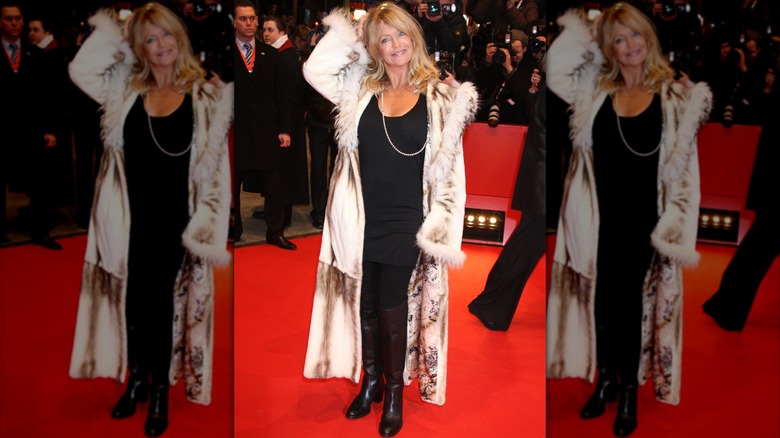 Goldie Hawn lors d'un événement sur le tapis rouge, vêtue d'une tenue entièrement noire et d'un long manteau soyeux avec une bordure en fourrure.