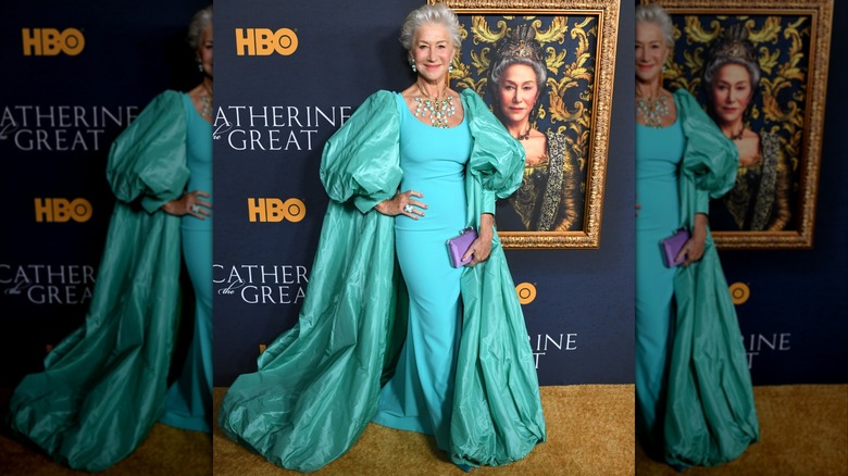 Helen Mirren posant devant un tableau en 2019 portant une robe turquoise avec une cape poofy et des bijoux tendance.
