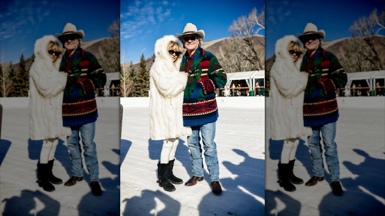 Goldie Hawn portant un manteau de fourrure blanc lie les bras avec Kurt Russell portant un pull multicolore et un jean