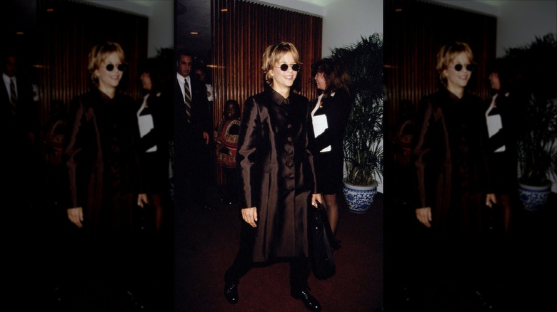Meg Ryan souriante et portant une robe-manteau marron et des lunettes de soleil.