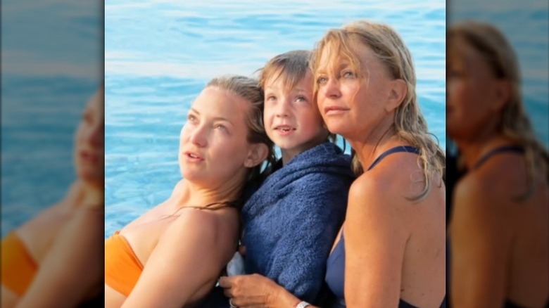 Kate Hudson, son fils Ryder et Goldie Hawn posent les yeux tournés vers le haut au bord de l'eau en maillot de bain.