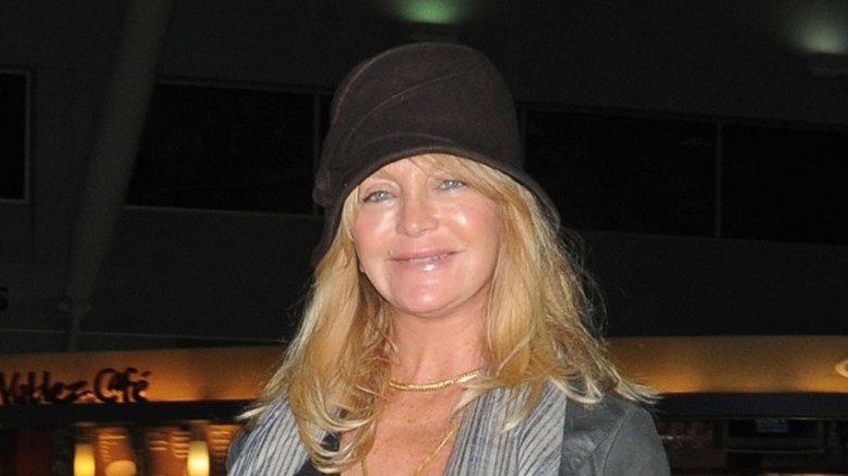 Goldie Hawn souriante portant un chapeau noir et une veste grise avec des colliers en or.
