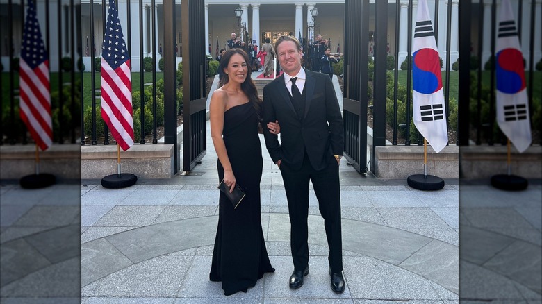 Chip Gaines en costume et Joanna Gaines vêtue d'une robe noire sans bretelles posant devant des drapeaux.