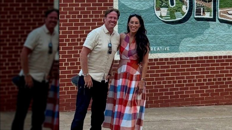 Chip Gaines dans une chemise boutonnée blanche et Joanna Gaines dans une robe à carreaux posant ensemble devant un mur de briques rouges.