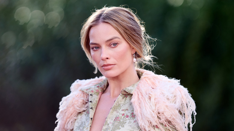 Margot Robbie posant dehors lors d'un événement, les cheveux relevés, vêtue d'une tenue détaillée et floue.