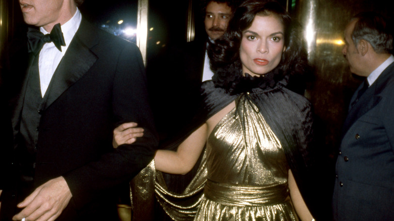 Bianca Jagger portant une robe dorée et une cape noire lors d'un événement nocturne.