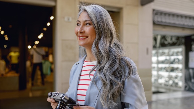 Une femme aux cheveux longs et ondulés souriante et tenant un appareil photo, vêtue d'une chemise rayée rouge et blanche et d'un blazer gris.