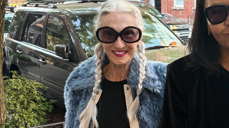 Une femme avec deux longues tresses souriant dans la rue, portant une veste en fourrure et de grandes lunettes de soleil.