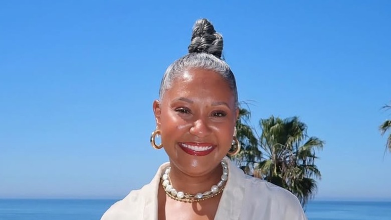 Une femme aux cheveux gris coiffés d'un chignon tressé portant une chemise blanche, des créoles et un collier tendance.