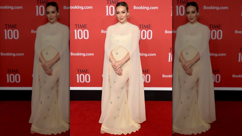 Hilary Duff posant avec ses mains devant elle lors d'un événement sur le tapis rouge dans une robe de couleur crème avec une cape.