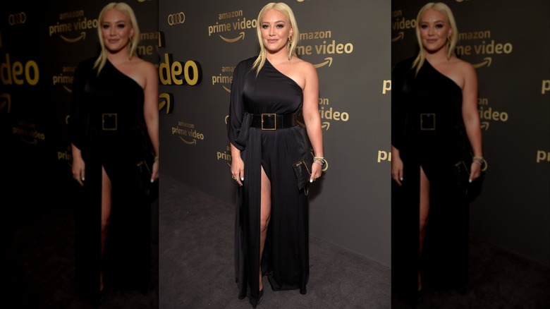 Hilary Duff souriant lors d'un événement sur le tapis rouge, vêtue d'une robe noire asymétrique avec une grande ceinture.