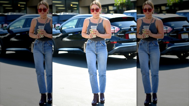 Hilary Duff photographiée dans la rue tenant une tasse et portant un débardeur gris, un jean, des bottes noires et des lunettes de soleil rouges.