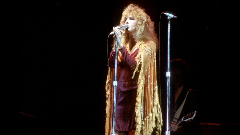 Stevie Nicks portant une tenue rouge avec une cape dorée chantant dans un micro avec Fleetwood Mac en 1977.