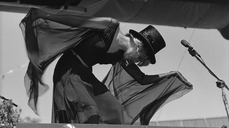 Photo en noir et blanc de Stevie Nicks portant une tenue noire fluide avec un haut-de-forme noir sur scène avec Fleetwood Mac en 1977.
