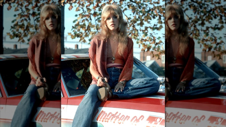 Stevie Nicks posant sur une voiture portant un jean et un haut rouge avec des chaussures à plateforme et une frange de rideau.