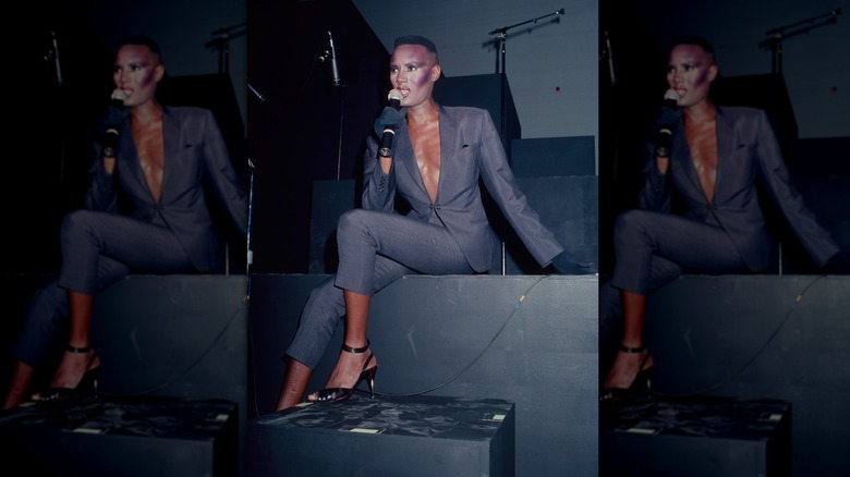 Grace Jones porte un costume gris avec un col en V profond alors qu'elle est assise et chante sur scène.