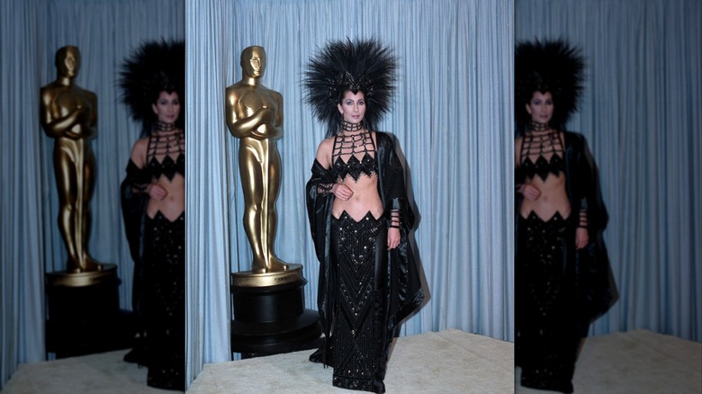 Cher portant une tenue noire pailletée et une grande coiffe noire à plumes devant une statue de récompense aux Oscars de 1986.