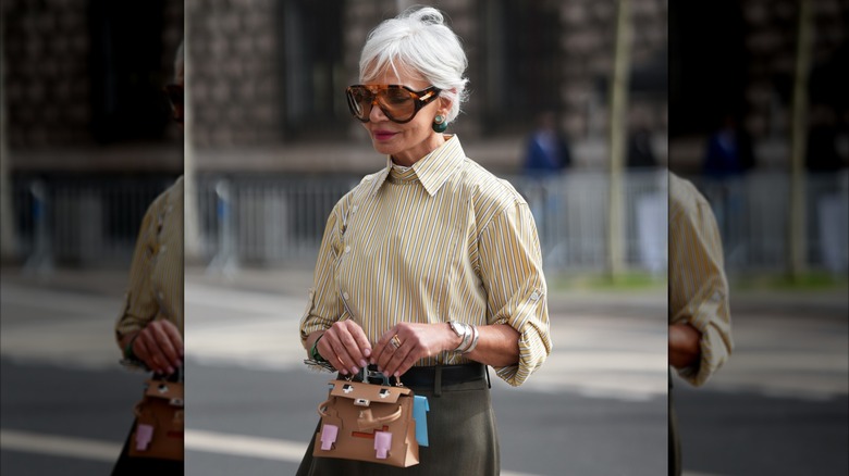 Une femme aux cheveux gris courts portant une chemise à col rayé et des lunettes de soleil avec des bagues en or et en argent et un bracelet en argent.