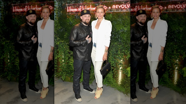 Benji Madden portant une tenue et un chapeau entièrement noirs posant devant un fond feuillu avec Cameron Diaz portant un look entièrement blanc.
