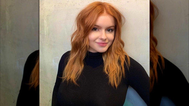 Ariel Winter posant dans un col roulé noir avec des cheveux roux ondulés sur un fond clair.