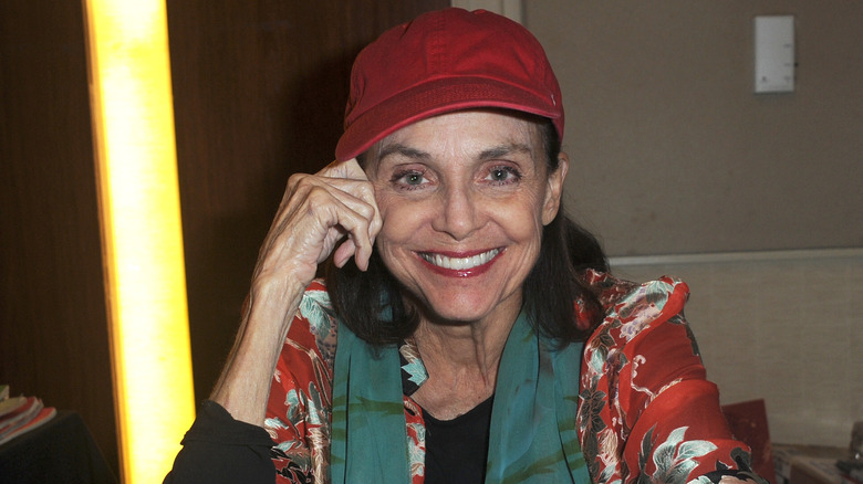 Valerie Harper assise à une table souriante et portant un chapeau rouge.
