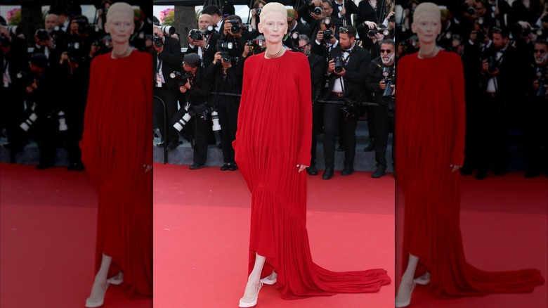 Tilda Swinton posant sur le tapis rouge avec son pied pointé et regardant sur le côté, vêtue d'une robe rouge fluide.