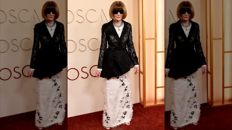 Anna Wintour sur le tapis rouge vêtue d'une robe blanche et noire duveteuse avec un manteau en dentelle noire, un collier tendance et des lunettes de soleil noires.