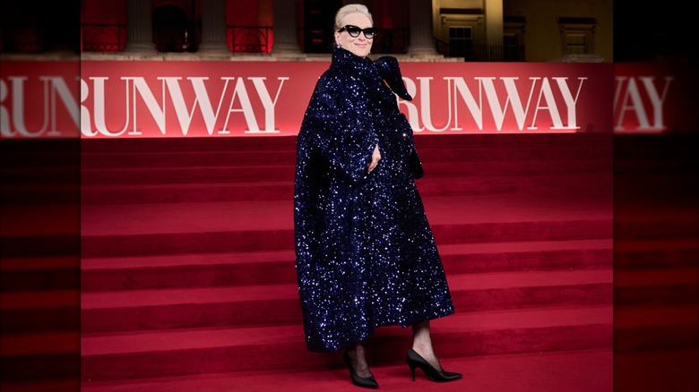 Meryl Streep sur le tapis rouge portant un manteau surdimensionné bleu foncé scintillant avec de simples escarpins noirs et des lunettes de soleil.