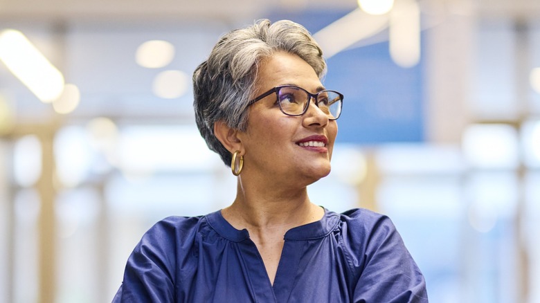 Une femme avec une coupe de lutin gris foncé portant des lunettes et un haut bleu regardant sur le côté.