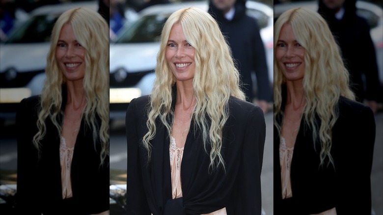 Claudia Schiffer souriante dans un haut blanc et un manteau noir avec des cheveux blonds en vagues de sirène.