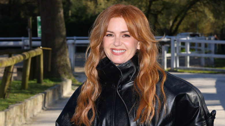 Isla Fisher souriante dehors, vêtue d'une veste en cuir noire, les cheveux détachés.