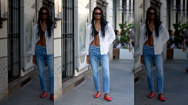 Une femme vêtue d'une chemise blanche, d'un jean et de ballerines rouges.