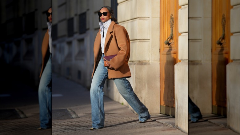 Une femme vêtue d'une chemise blanche, d'un jean et d'un manteau marron.