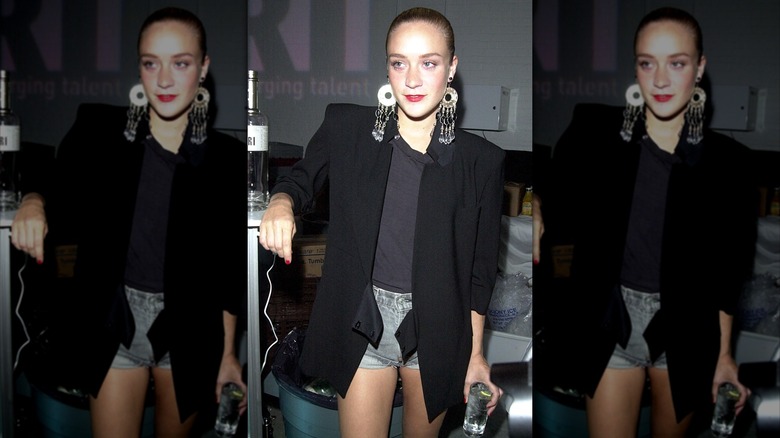 Chloë Sevigny photographiée avec un short en jean, un t-shirt noir, un blazer noir oversize et des boucles d'oreilles tendance avec les cheveux lissés en arrière.